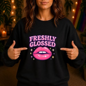 Könnte beinhalten: Schwarzes Sweatshirt mit dem Text "FRESHLY GLOSSED" in Pink und Lila, über einer rosa Lippen-Grafik. Das Design enthält kleine Sternakzente. Ein lässiger Stil.