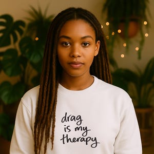 Könnte beinhalten: Cremefarbenes Sweatshirt mit dem Text "drag is my therapy" in schwarzer Schrift. Die Person im Bild hat lange, braune Dreadlocks. Der Hintergrund zeigt grüne Pflanzen und sanfte Beleuchtung.