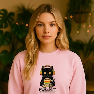 Könnte beinhalten: Hellrosa Sweatshirt mit einer Grafik einer schwarzen Katze mit Kopfhörern, die eine Tasse und einen Gamecontroller hält. Der Text "PAWS & PLAY" und "QUEER SERVED WARM" ist unter der Katzenillustration aufgedruckt.