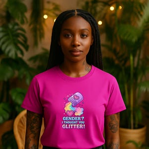 Könnte beinhalten: Ein magentafarbenes T-Shirt mit dem Text "Gender? I thought you glitter!" und einer Grafik einer Glitzerflasche, aus der ein Regenbogen quillt. Das T-Shirt wird von einer Person mit langen schwarzen Zöpfen getragen.