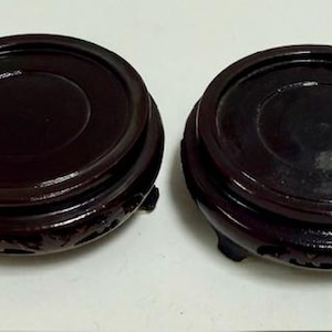 May include: Two dark brown, wooden display stands with a glossy finish. Each stand has a round top with a recessed center and a decorative carved design around the base. The stands are elevated by three small feet.