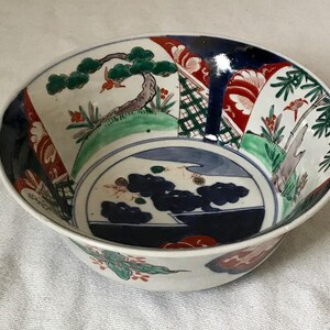 Op de afbeelding: Een witte keramische kom met een blauw, groen, rood en zwart bloemenontwerp. De kom heeft een brede rand en een afgeronde bodem.