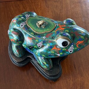 Peut inclure: Une figurine de grenouille en cloisonné vert avec une grenouille dorée sur le dessus. La grenouille est décorée de détails colorés et repose sur une base en bois.