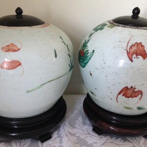 May include: Two white porcelain jars with lids and floral designs in red, green, and brown. The jars are on wooden stands.