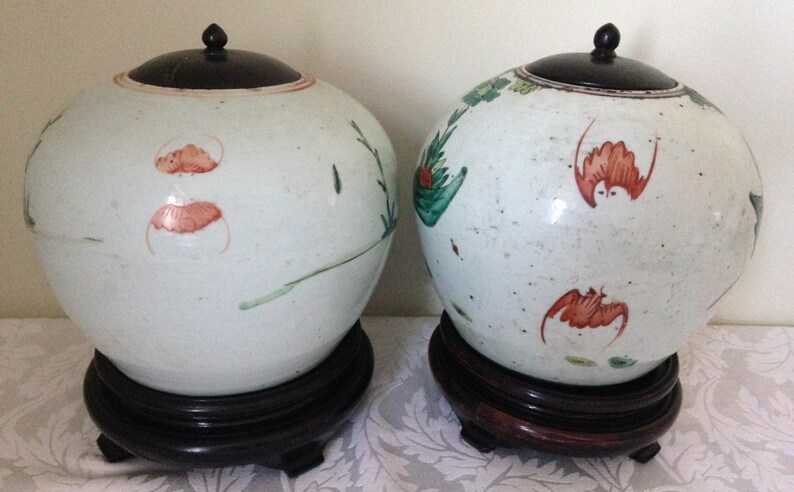 May include: Two white porcelain jars with lids and floral designs in red, green, and brown. The jars are on wooden stands.