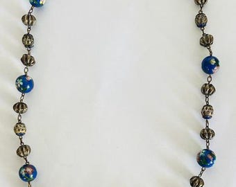 Puede incluir: Un collar vintage con cuentas florales azules alternadas con cuentas de metal ornamentadas. Las cuentas azules tienen un diseño floral en rosa, amarillo y blanco. Las cuentas de metal son de color bronce. Una pieza de joyería clásica.