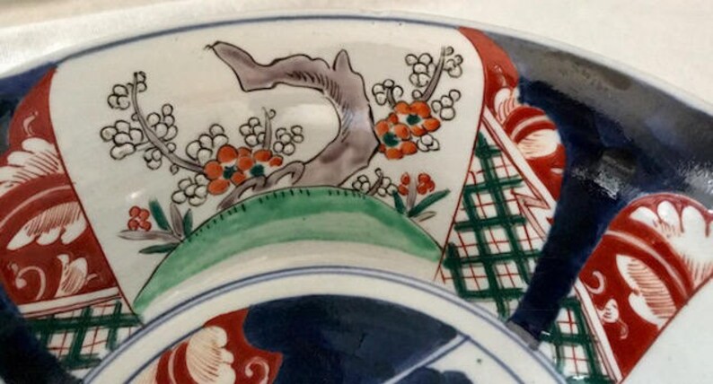 Op de afbeelding: Een grote, witte keramische plaat met een blauw, groen en rood bloemenontwerp. De plaat heeft een gestileerde boom met rode bloemen en groene bladeren, evenals een geometrisch patroon op de achtergrond.