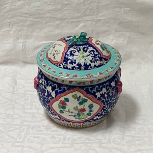 Puede incluir: Un tarro de porcelana azul y blanco con tapa, con diseños florales y geométricos. El tarro tiene dos asas rosas y un pomo verde en la tapa.