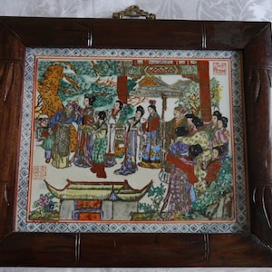 May include: A framed porcelain tile with a colorful scene of people in traditional Chinese clothing. The tile is surrounded by a dark wood frame with a decorative design.