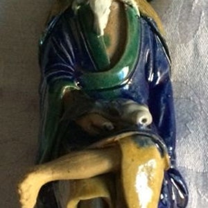 May include: A ceramic figurine of a man wearing a green hat and blue robe. He is sitting with his legs crossed and holding a small object in his hand.