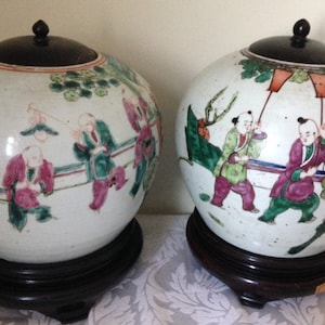 May include: Two white porcelain lidded jars with black wooden lids and bases. The jars are decorated with colourful hand-painted scenes of people and landscapes.