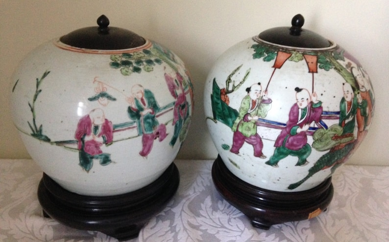 May include: Two white porcelain lidded jars with black wooden lids and bases. The jars are decorated with colourful hand-painted scenes of people and landscapes.