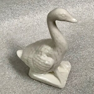 Puede incluir: Figurita de ganso de cerámica blanca con un cuerpo texturizado detallado y un cuello largo. El ganso está de pie sobre una base cuadrada.