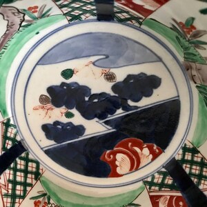 Op de afbeelding: Een wit keramiek bord met een blauw en wit landschapsscene geschilderd op de bodem. De sc&egrave;ne omvat een bergketen, een rivier en een paar bomen. Het bord is versierd met een rood en wit bloemenontwerp rond de rand.