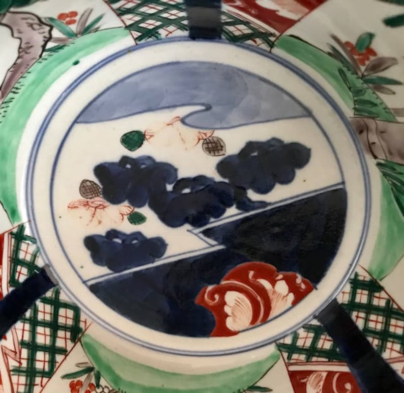 Op de afbeelding: Een wit keramiek bord met een blauw en wit landschapsscene geschilderd op de bodem. De sc&egrave;ne omvat een bergketen, een rivier en een paar bomen. Het bord is versierd met een rood en wit bloemenontwerp rond de rand.