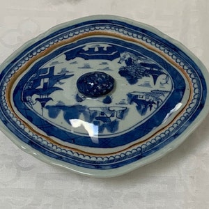 May include: A blue and white porcelain lidded dish with a scalloped edge and a knob on the top. The dish is decorated with a Chinese landscape scene in blue and white.