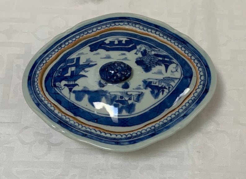 May include: A blue and white porcelain lidded dish with a scalloped edge and a knob on the top. The dish is decorated with a Chinese landscape scene in blue and white.