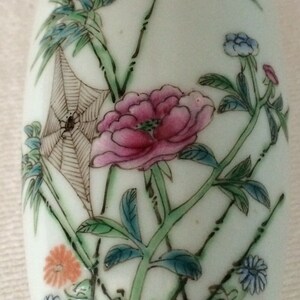 Może przedstawiać: Biały ceramiczny wazon z kwiatowym wzorem. Wazon przedstawia r&oacute;żowe kwiaty, zielone liście i pajęczynę z pająkiem w środku. Wazon jest ozdobiony jasnoniebieskim i zielonym schematem kolorystycznym.