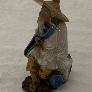 Op de afbeelding: Een keramiek beeldje van een zittende man met een brede hoed en een lange robe. Hij heeft een lange witte baard en houdt een pijp in zijn hand. Het beeldje is geschilderd in tinten bruin, wit en blauw.