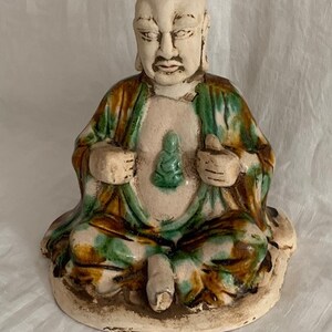 Peut inclure: Une statue en céramique d'un Bouddha assis avec un glacis vert et brun. Le Bouddha tient un plus petit Bouddha vert sur ses genoux.