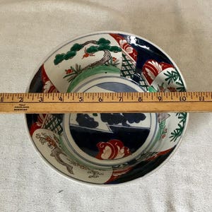 Op de afbeelding: Een witte keramische kom met een blauw, groen, rood en zwart geverfd ontwerp. De kom heeft een landschapssc&egrave;ne met een dennenboom, bergen en een rivier. De kom heeft een diameter van ongeveer 23 cm.