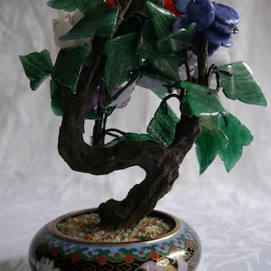 May include: A jade bonsai tree with colourful gemstone flowers in a black and gold cloisonné pot. The flowers are made of amethyst, rose quartz, lapis lazuli, and other gemstones.
