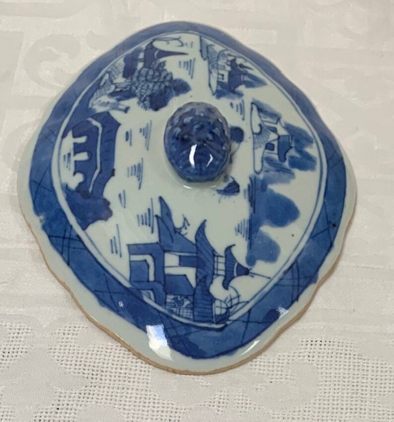 May include: A blue and white porcelain lid with a scalloped edge and a knob on top. The lid features a Chinese landscape design with buildings and trees.