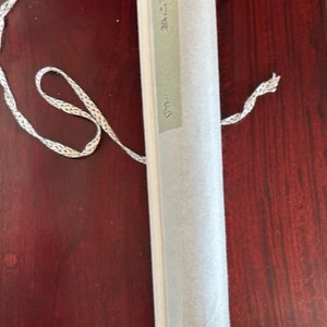 May include: A rolled-up scroll with a white paper exterior and a black base. A white and silver cord is attached to the scroll. The scroll has a rectangular label with Japanese text.