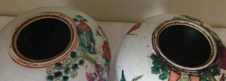 May include: Two white ceramic jars with lids, decorated with floral and figural designs in shades of green, red, and orange. The jars have a black interior.