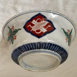 Op de afbeelding: Een witte keramische kom met blauwe en rode beschilderde ontwerpen. De kom heeft een centraal ruitvormig ontwerp met een rode achtergrond en een blauwe rand. De kom heeft ook bloemmotieven in groen, rood en blauw.