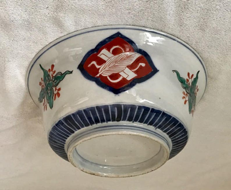 Op de afbeelding: Een witte keramische kom met blauwe en rode beschilderde ontwerpen. De kom heeft een centraal ruitvormig ontwerp met een rode achtergrond en een blauwe rand. De kom heeft ook bloemmotieven in groen, rood en blauw.
