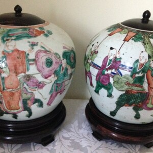 May include: Two white porcelain Chinese ginger jars with lids and painted with colourful figures and floral designs. The jars are on wooden stands.