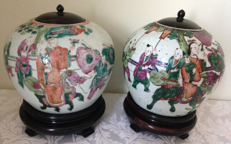 May include: Two white porcelain Chinese ginger jars with lids and painted with colourful figures and floral designs. The jars are on wooden stands.