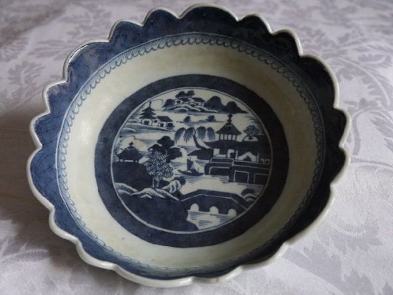 K&ouml;nnte beinhalten: Eine wei&szlig;e Porzellanschale mit einem gewellten Rand und einem blauen und wei&szlig;en Landschaftsdesign. Die Schale zeigt eine Br&uuml;cke, B&auml;ume und Geb&auml;ude.