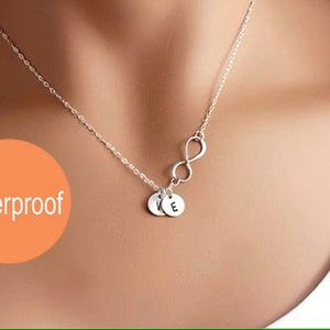 May include: A silver infinity necklace with two small silver circles hanging from it. The circles have the letters "V" and "E" engraved on them. The necklace is on a model's neck. The word "Waterproof" is written in orange on a peach background.