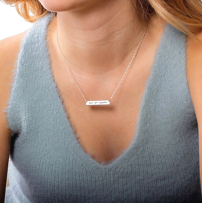 May include: A silver-toned horizontal bar necklace featuring a delicate chain and a rectangular pendant engraved with the cursive text 'love you mom'. The metal bar is polished and suspended centrally from a thin, matching silver-colored link chain.