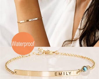 May include: A gold-toned metal bracelet featuring a thin, flat horizontal bar engraved with the name EMILY in capital letters. The bracelet includes a delicate chain, a small blue circular accent stone, and a lobster claw clasp closure.