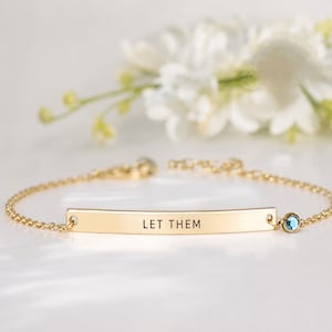 May include: A gold-toned chain bracelet featuring a central curved metal bar engraved with the text "LET THEM" in black capital letters. A small, round, light blue gemstone is set in a gold-toned bezel attached to the right side of the bar.