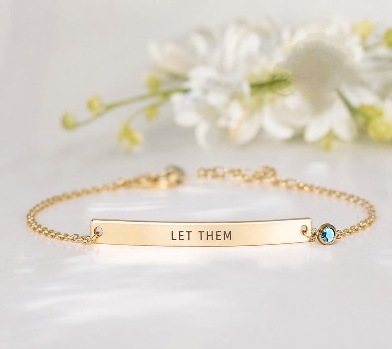May include: A gold-toned chain bracelet featuring a central curved metal bar engraved with the text "LET THEM" in black capital letters. A small, round, light blue gemstone is set in a gold-toned bezel attached to the right side of the bar.
