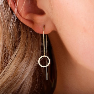 May include: Gold chain earrings with a small circle pendant and a thin, straight bar hanging below. The earrings are shown on a person's ear.