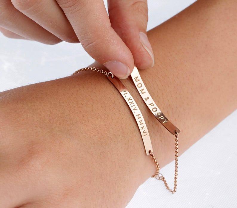 May include: Two rose gold-toned bar bracelets with delicate chain links. One bar is engraved with the text MOM & POPPY, while the other bar features the Roman numeral date VI.XXIV.MMXVI. The bracelets are worn together on the wrist.