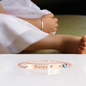 May include: A rose gold bracelet with a personalised nameplate that reads "Daisy" with a heart symbol. The bracelet has a small blue gemstone charm.