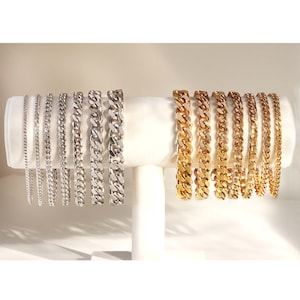 May include: A display of silver and gold curb chain bracelets of varying widths. The bracelets are arranged in a row on a white display stand.