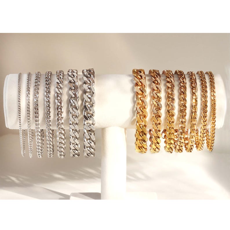 May include: A display of silver and gold curb chain bracelets of varying widths. The bracelets are arranged in a row on a white display stand.