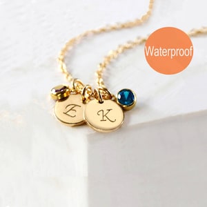 May include: Gold necklace with two round charms engraved with the letters "E" and "K". The necklace also features two small gemstones, one purple and one blue. The text "Waterproof" is visible in the image.