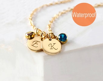 May include: A gold-toned necklace featuring two circular charms engraved with the letters E and K, accompanied by a purple and a blue gemstone charm. An orange circular graphic with the text Waterproof is visible in the upper right corner.