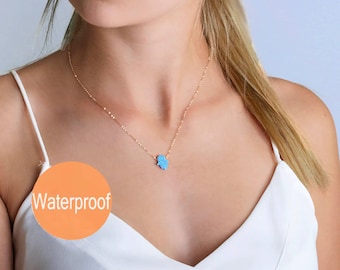 May include: A delicate gold-tone chain necklace featuring a small, light blue hamsa hand pendant with an iridescent finish. The necklace is displayed against a white top and includes an orange circular graphic with the text Waterproof.