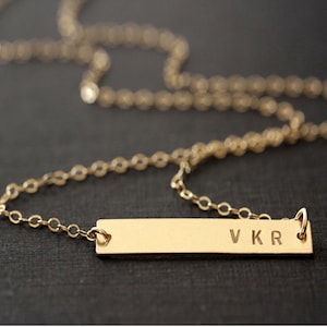May include: A gold-toned bar necklace featuring a delicate chain and a rectangular pendant. The pendant is engraved with the capital letters V K R on the right side. The metal has a smooth, polished finish, and the chain consists of small circular links.