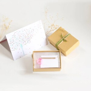May include: A gold-toned bar necklace rests inside a small rectangular cardboard gift box with a clear lid, accompanied by a pink flower accent. A matching cardboard gift box with green ribbon and a greeting card featuring a heart tree are included.