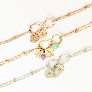 May include: Three gold infinity symbol necklaces with small charms. The charms are personalized with the letters R, A, K, S, J, M, and E. The necklaces are layered on a white surface.